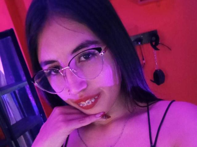 MiaDiiaz with purple-tinted hair, glasses, and red lipstick, biting her finger, showing her teeth, wearing a black strappy top, and a silver necklace.