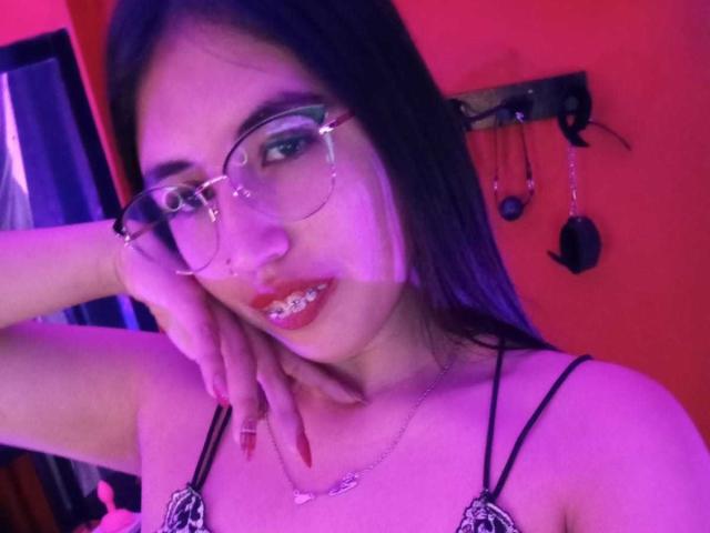 MiaDiiaz with purple-tinted skin, black hair, glasses, and red lipstick, showing metal teeth, wearing a black strappy top, hand on cheek, and a necklace.