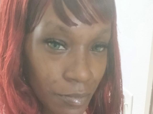 A Prettycat87 tem pele marrom escura, olhos verdes e cabelo ruivo com franja. Seus lábios brilhantes e sorriso discreto são visíveis. Seu rosto apresenta algumas pequenas imperfeições..