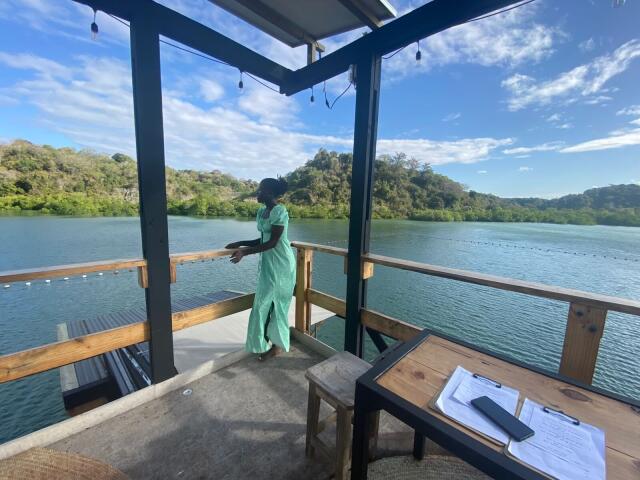 A photograph of KharlaQueen standing on a wooden deck by a lake, wearing a green jumpsuit. She holds a black object in her right hand and looks out at the water. The deck has a wooden table with a clipboard and papers on it. The clear blue sky and green hills are in the background. Bright sunlight casts shadows. KharlaQueen is in the center, with the lake and hills to the right.