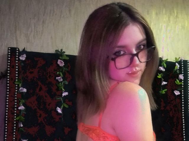 ZoeyClark with glasses, looking over her shoulder, wearing an orange lace bra, showing her small tits, and a tattoo on her right shoulder. Brown hair, slight pout.