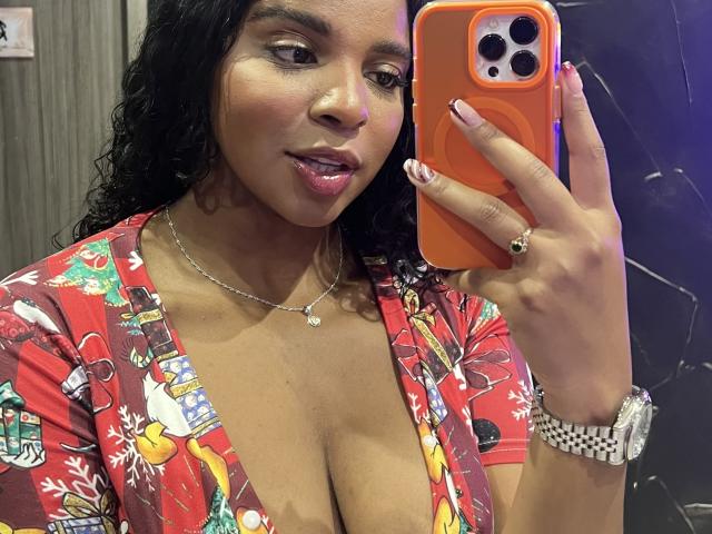 LisaMendez with curly black hair, brown skin, and pink lipstick, wearing a red floral top with deep cleavage, holding an orange phone, showing her boobs, and a silver watch.