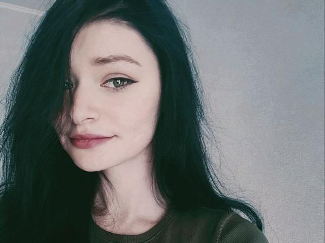 SweetNalya with pale skin, dark black hair, green eyes, and a subtle smile, wearing a green shirt, has a small nose and full lips, looking slightly to the left.