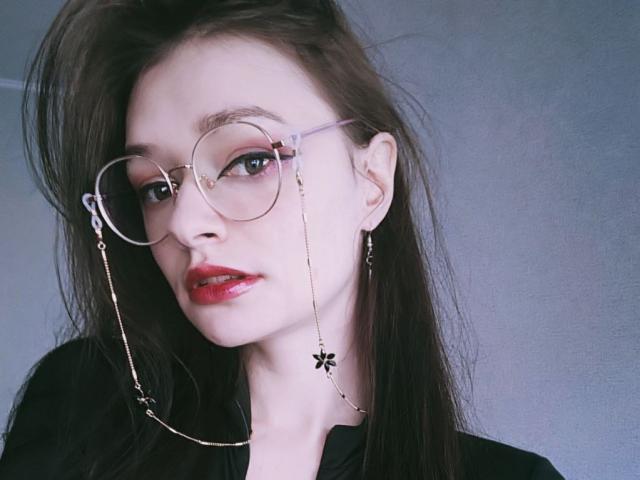 SweetNalya with round glasses, red lipstick, black hair, and gold chain earrings, showing her white face and black shirt, looking slightly to the side with a confident expression.