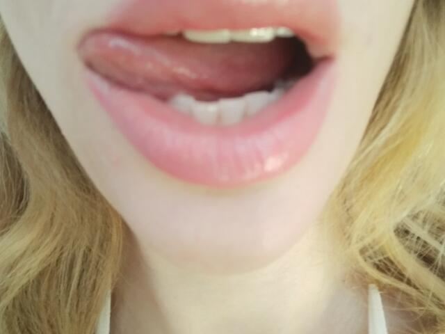 Close-up of MissFrenchy's face showing her open mouth with her tongue slightly out. She has pink glossy lips and white teeth. Her blonde hair is on either side of her face. The image focuses on her lips and tongue, with her skin appearing fair. The texture of her lips is smooth and shiny. The composition is centered on her mouth.