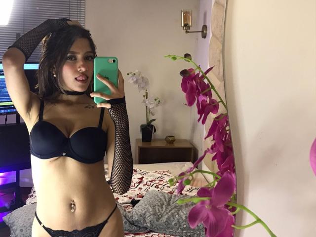 AfroditaWild trägt einen schwarzen BH und Unterhose, Netzhandschuhe, eine Halskette und lila Lippenstift, hält ein grünes Telefon und macht ein Selfie vor einem Spiegel, in dem eine rosa Orchidee zu sehen ist. Brüste, Nippel, Vulva sind sichtbar..
