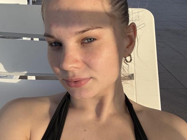 SummerSaisa has light skin, blue eyes, and brown hair in a bun. She's wearing a black halter top, small hoop earrings, and sitting outside in sunlight.