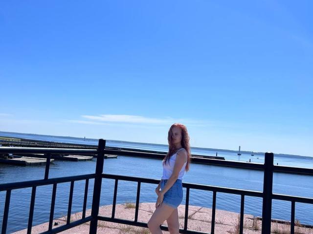 SummerSaisa with long red hair, white tank top, and denim shorts, standing by a black railing, blue water, and clear sky, looking at camera, sunny day.