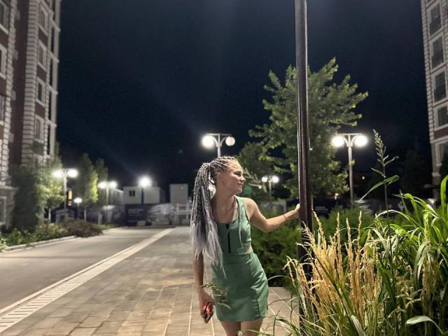 SummerSaisa with silver braided hair, green dress, standing by a lamp, holding red phone, night, sidewalk, grass, streetlights, tall buildings, looking left, urban setting, casual.