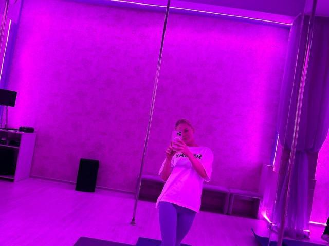 SummerSaisa with blonde hair, wearing a white tee and blue leggings, holding a phone, standing in a purple-lit room with a pole and mirror.