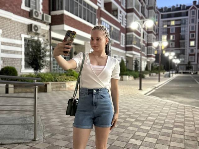 SummerSaisa with braided hair, white top, and blue shorts, holding phone, taking selfie, black crossbody bag, standing on tiled sidewalk, evening, streetlights, buildings in background.