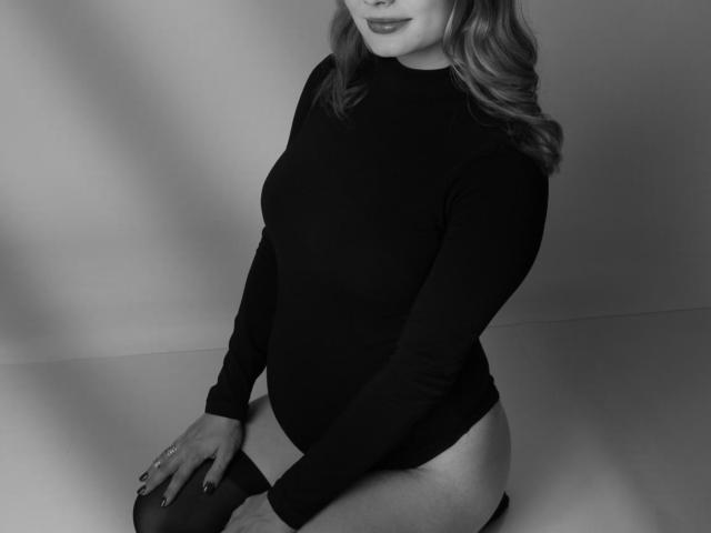 SummerSaisa in a black long-sleeve leotard, showing her ass, sitting with knees up, wavy blonde hair, subtle smile, gray-black photo, hands on thighs, nails painted dark.
