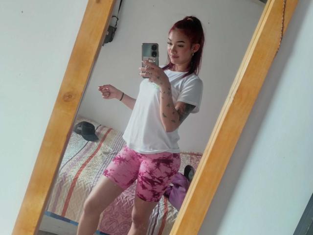 SashaHans with red ponytail, white tee, pink floral shorts, tattoos, holding phone, mirror selfie, standing, wooden frame, bed with striped blanket in background.