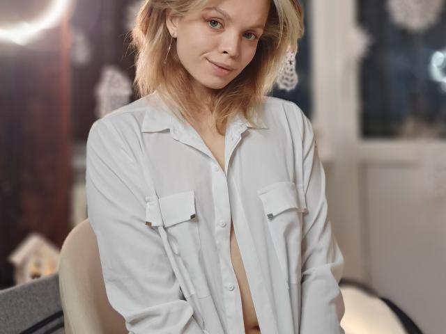 Sinteas has blonde hair, light skin, and small tits, wearing an unbuttoned white shirt, smiling slightly, with a subtle, confident, sexy look.