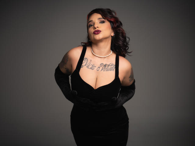 Alexandrapsique, avec de grandes seins, une robe noire, une poitrine tatouée, des gants noirs, un collier de perles, des cheveux noirs, une pose confiante, fond gris..