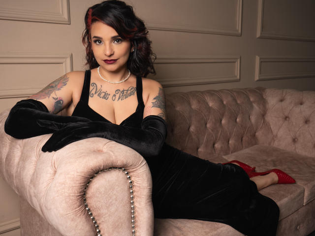 Alexandrapsique, com tatuagens, vestindo um vestido preto e luvas, deitada em um sofá de veludo rosa, usando saltos vermelhos e um colar de pérolas, mostrando seus seios e com um sorriso confiante..
