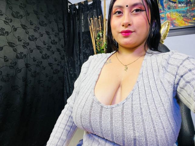 KalpanaAisha with large boobs, wearing a grey ribbed top, pink lipstick, black hair, small tattoo on left shoulder, gold necklace, sitting in a chair, smiling.