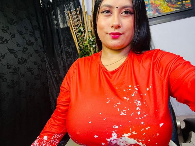 KalpanaAisha with large tits, wearing a red shirt with white splashes, black hair, pink lipstick, and a small bindi on her forehead, sitting in a chair.