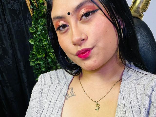 KalpanaAisha with black hair, pink lipstick, nose ring, and small dot on forehead. Wearing gray sweater, gold necklace, hoop earrings, and has tattoo on chest. Smiling slightly.