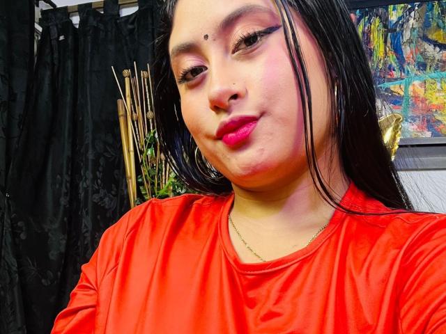 KalpanaAisha with dark straight hair, red shirt, pink lipstick, black eyeliner, small mole above her nose, and a slight smirk, wearing a gold necklace, against a black curtain.