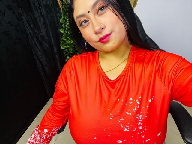 KalpanaAisha with big tits in a shiny red top, black hair, pink lipstick, and a small dot on her forehead, sitting and smiling. Background is mostly black.