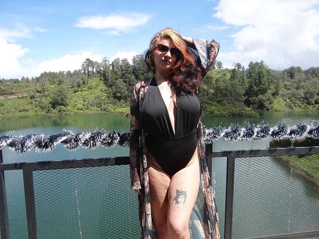 Alexandrapsique with big tits in a black one-piece, open robe, wearing sunglasses, standing on a balcony with a tattoo on her thigh, by a green water body.