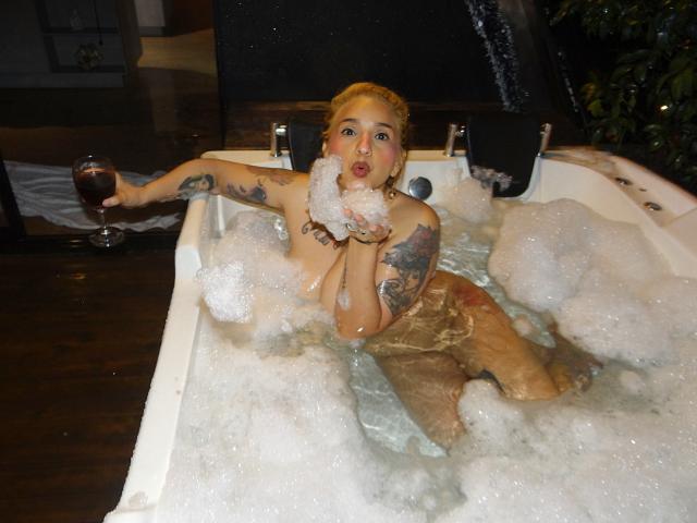 Alexandrapsique with tattoos, naked, in a bubbling hot tub, holding a glass of red wine, foam on her tits and arm, looking surprised with her mouth open.