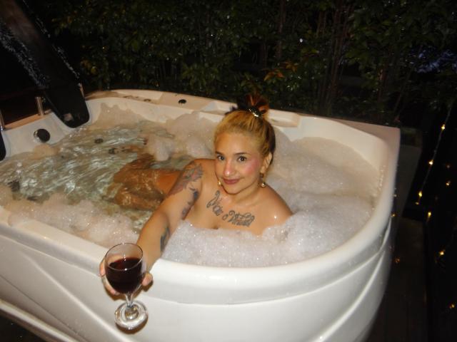 Alexandrapsique with blonde hair in a bun, tattoos on chest and arm, naked in a bubbling white hot tub, holding a glass of red wine, smiling.