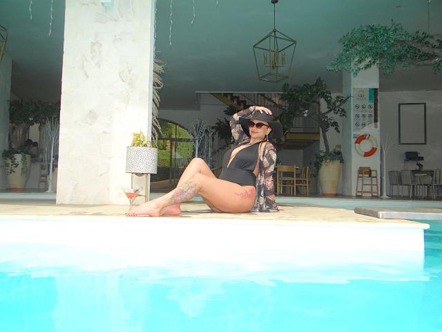Alexandrapsique, sitting by the pool, wearing a black one-piece, showing tattoos on arms and legs, with a sunhat, sunglasses, and a visible ass. Blue water in foreground.