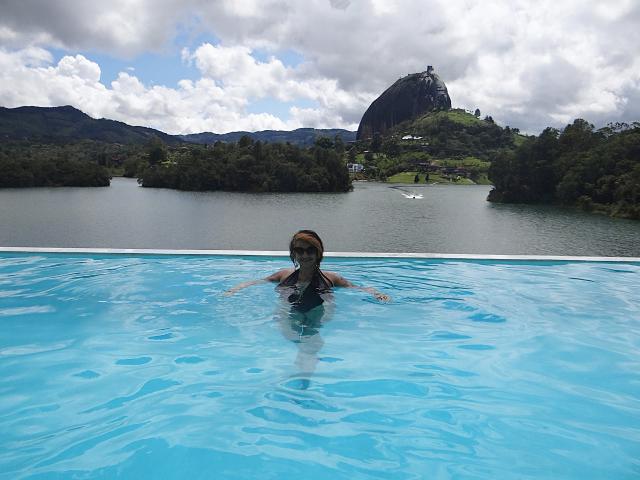 Alexandrapsique's blue-tinted tits are barely above the water, wearing dark sunglasses, in a clear pool with a mountain in the background under a cloudy sky.