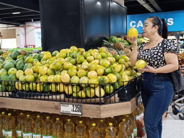 Amarantac with yellow-green and green lemons, holding one lemon, wearing a black and white polka dot top, and blue jeans, standing in front of a wooden display.