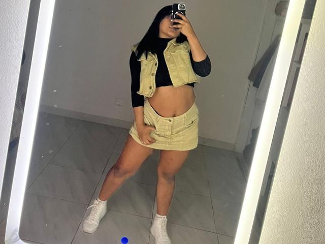 Sarita, un look mignon avec un ventre légèrement visible, un haut noir à manches longues, un gilet jaune court, une mini-jupe jaune, des baskets blanches, tenant un téléphone, debout devant un miroir, cheveux foncés, peau bronzée..