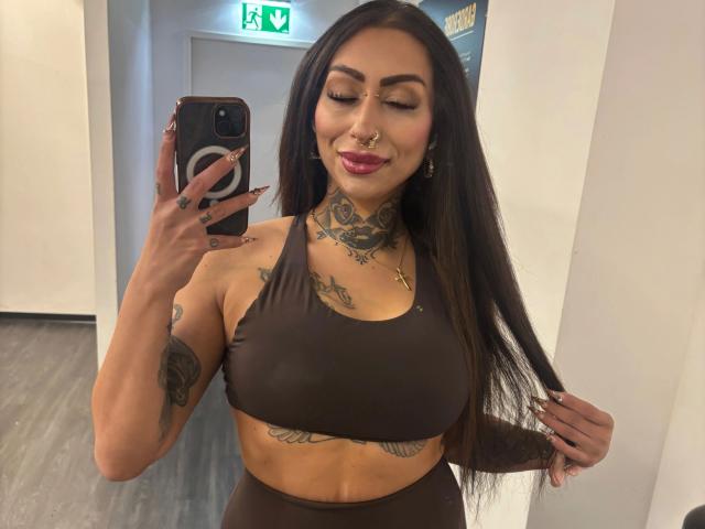 Pinkerbell-hot with long black hair, brown sports bra, tattoos, nose ring, and pink lipstick, taking a mirror selfie with her phone, holding her hair, smiling. Boobs visible.