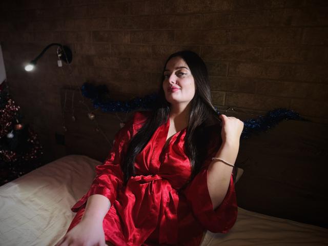MilanaPrice with long dark hair, red satin robe, sitting on bed, hand in hair, smiling, soft light, Christmas tree with red ornaments on left. Her tits are visible.