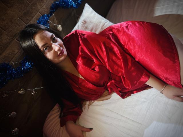 MilanaPrice with long black hair, red silky robe, lying on white bed, looking up with a slightly pouty face, showing her tits and ass. Blue tinsel in background.
