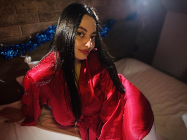 MilanaPrice with long black hair, wearing a shiny red silk robe, leaning on a bed, showcasing her big tits and a cute smile, in a dimly lit room.