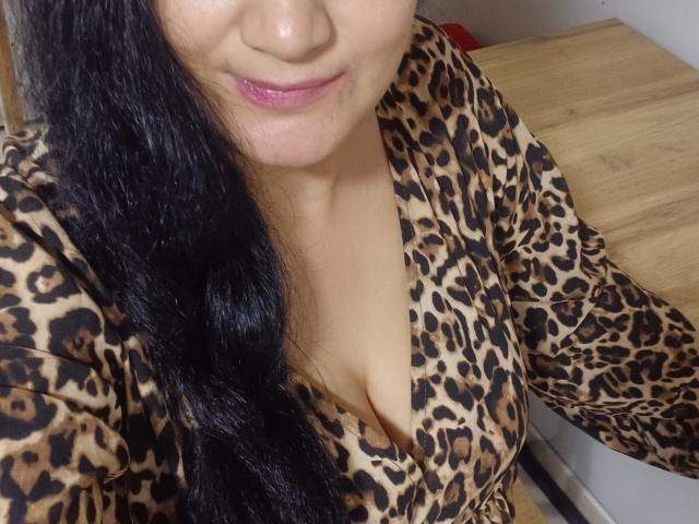 KarinaCecillia, upper body shot, shows a close-up of her face and chest. She has long, wavy black hair and light brown skin. She wears a leopard-print dress with a deep V-neck, revealing cleavage. Her lips are slightly parted, showing pink lipstick. The dress's pattern is bold with brown spots on a beige background. The wooden table in the background is blurred. The image has a casual, selfie-style composition.