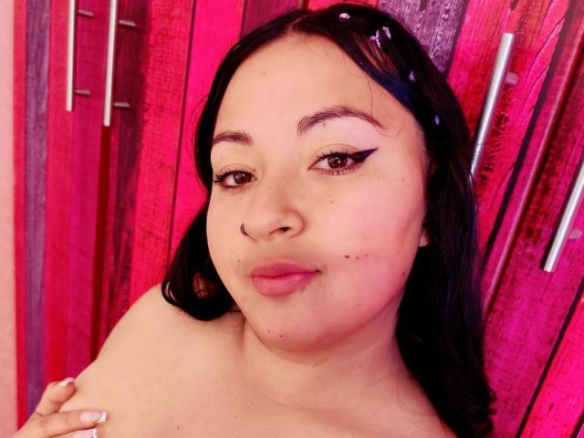 LindaGigi with dark hair, pink lipstick, and eyeliner, showing a small pimple on her left cheek, against a pink wooden background. She's topless, with a black hair clip.