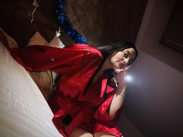 MilanaPrice in a red satin robe, sitting on a bed, holding a flashlight to her face, with long dark hair, soft light, and a sexy, relaxed expression.