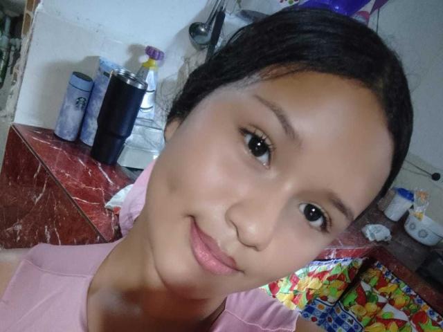 MiaCelestee's close-up selfie shows a young woman with smooth, light brown skin and dark, straight hair pulled back. She has small, dark eyes and a subtle, calm smile. She wears a pink shirt. The background includes a red marble countertop with a spray bottle, a can, and a colorful, patterned object. The composition focuses on her face, with a slight tilt to her head. The lighting is even, highlighting her clear skin and relaxed expression.