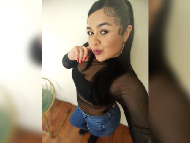 HannahMolly, avec de longs cheveux noirs, portant un haut noir transparent, un jean bleu et des ongles rouges, pointant vers la caméra, souriant, avec des yeux marron, des boucles d'oreilles, un sol en bois et une chaise verte..