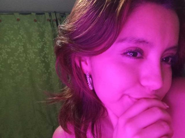 LolaBubbles with pink-tinted skin, brown wavy hair, and pink earrings, biting her finger, looking slightly to the side with a subtle smile. Her tits are partially visible.