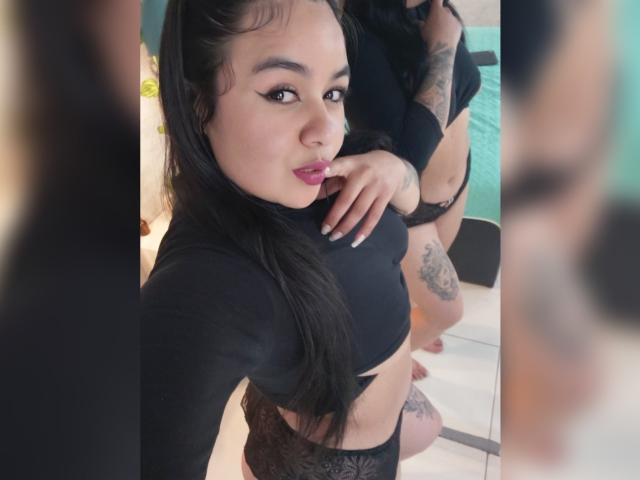 HannahMolly with black hair, wearing a black crop top and lace panties, has her finger to her lips, showing tattoos on her arm and thigh, in a mirror selfie.