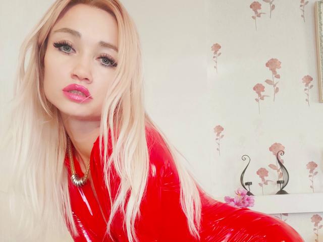 AlinaFit with blonde hair, red glossy latex, pink lipstick, and thick eyelashes, wearing a gold necklace, looking directly at the camera with a slight pout.
