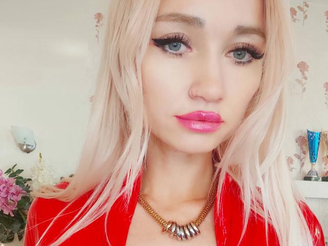 AlinaFit with blonde hair, blue eyes, and glossy pink lips. She's wearing a red shirt, gold necklace with metallic spikes, and has thick black eyeliner. Boob shot, face forward.