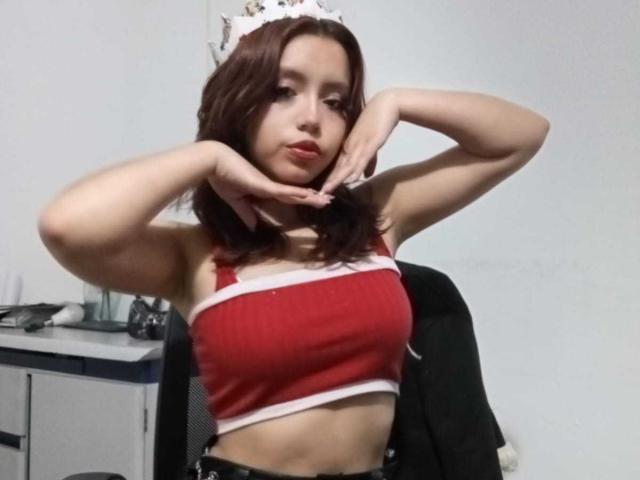LolaBubbles with brown hair, wearing a red crop top, white trim, and a white headpiece, posing with hands near her face, showing her small tits and a cute face.