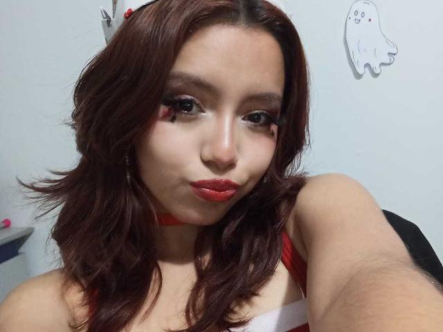 LolaBubbles with red lipstick, dark wavy hair, and red choker, pouting slightly, wearing a red and white top, showing her tits, in a selfie.