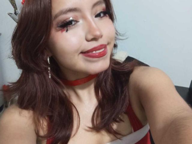 LolaBubbles has red lipstick, long brown hair, and a red choker. She's smiling with cleavage visible, wearing red makeup and earrings. She has a cute, sexy look.