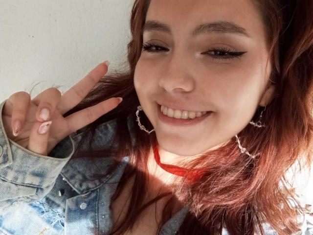 LolaBubbles, with red hair, wearing a denim jacket, and red choker, makes a peace sign, smiling with winged eyeliner, and cute earrings, in a bright, sunlit selfie.