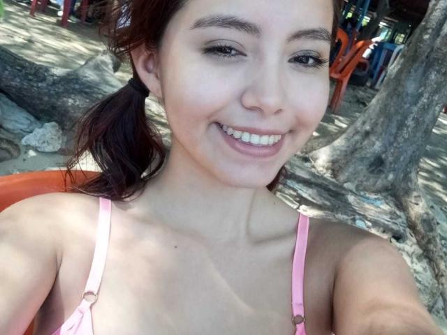 LolaBubbles with light skin, brown eyes, and a smile, wearing a pink bikini top, showing small tits, and red pigtails, sitting near a tree on a sandy beach.